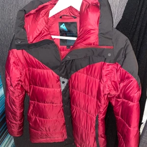 Röd och svart pufferjacka  - Säljer en snygg röd och svart pufferjacka med huva. Jackan har en dragkedja och knappar framtill för extra skydd mot kyla. Perfekt för kalla dagar med sin vadderade design.  Märket är klättermussen