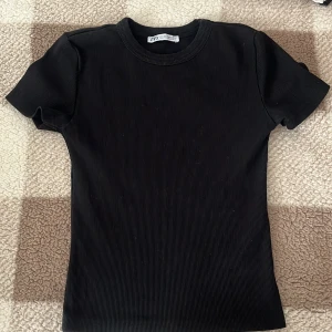 Svart ribbad t-shirt från Zara - Säljer en stilren svart ribbad t-shirt från Zara. Den har en klassisk rund hals och korta ärmar, perfekt för en enkel och snygg look. Passar bra till både jeans och kjol. 🖤