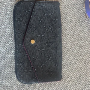 Svart kuvertväska med monogram - Elegant svart kuvertväska med präglat monogrammönster och guldfärgad knapp. Väskan har en kedjerem och insidan är klädd i vinrött tyg. Perfekt för att bära det nödvändigaste med stil. Ge din outfit en elegant toch!