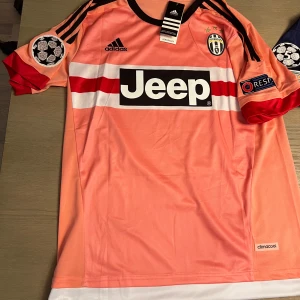 Rosa Juventus fotbollströja med Pogba 10 - Säljer en rosa Juventus fotbollströja från Adidas med Pogba och nummer 10 på ryggen. Tröjan har korta ärmar och ett svart Jeep-tryck på framsidan. Den har även svarta och röda detaljer samt Juventus-emblem på bröstet.