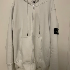 Vit zip hoodie från Stone Island - Säljer en stilren vit zip hoodie från Stone Island med dragkedja och klassisk logga på ärmen. Finns en liten fläck (bild 4 visar) men den är knappt synlig på håll. Annars bra skick. 