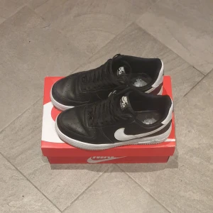 Svarta Nike Air Force 1 - Säljer ett par klassiska svarta Nike Air Force 1 sneakers. De har en stilren design med svart skinn och vit sula. Skorna har den ikoniska Nike-loggan i vitt på sidan och 'Air' texten på hälen. Perfekta för både vardag och fest!