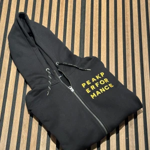 Svart hoodie från Peak Performance - Säljer en snygg svart hoodie från Peak Performance. Den har en dragkedja framtill och text i gult på bröstet. Perfekt för en casual look eller träning. Den är i bra skick och superbekväm!