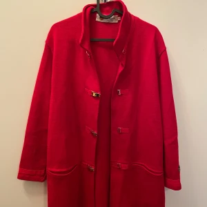 Röd kappa från Busnel - Säljer en elegant röd kappa från Busnel, tillverkad i Frankrike. Den har en klassisk design med långa ärmar och dekorativa knappar framtill. Perfekt för att ge en färgklick till din höst- och vintergarderob. Kappan är i storlek 38 och har en tidlös stil som passar till många olika tillfällen.