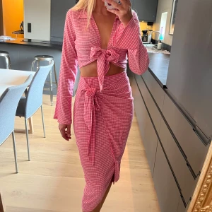 Rosa Set - Ett rosa och vit randigt set med kjol och topp.  Kjolen har resår i midjan och knyts på sidan. Midi lång.  Toppen är långärmad med krage och utsvängda ärmar. Den knyts framtill. Något croppad.  Superfint set, perfekt för den som gillar färg! Strl S