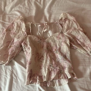 Blommig topp från H&M - Säljer en superfin blommig topp från H&M i storlek XS. Den har en romantisk stil med puffiga, långa ärmar och en söt volangdetalj längst ner. Toppen är i en ljusrosa nyans med blommönster och har en smockad, tight passform. Perfekt för vår och sommar!