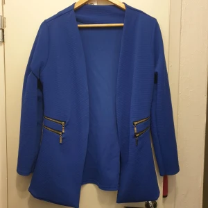 Blå kavaj med dragkedjedetaljer - Säljer en snygg blå kavaj med coola dragkedjedetaljer på fickorna. Den har en stilren design utan knappar och är perfekt för både vardag och fest. Kavajen är långärmad och har en skön passform.  Passar S o M. 