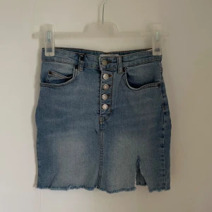 Jeanskjol från Pull&Bear - Säljer en snygg jeanskjol från Pull&Bear i storlek S. Kjolen är i en klassisk blå denimfärg och har en cool knappdetalj framtill. Den har en rå kant nertill och en liten slits som ger den en avslappnad look. Perfekt för både vardag och fest!
