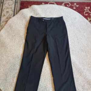 Svarta kostymbyxor från Batistini - Säljer ett par stiliga svarta kostymbyxor från Batistini. De är gjorda av en skön blandning av ull och polyester med lite lycra för stretch. Byxorna har en klassisk rak passform och är perfekta för både formella tillfällen och kontoren.