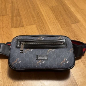 Gucci midjeväska med tigertryck - Säljer en snygg Gucci midjeväska i svart med coolt tigertryck. Väskan har en dragkedja framtill och ett justerbart band i rött och blått. Perfekt för att hålla dina saker säkra och stiliga på samma gång!
