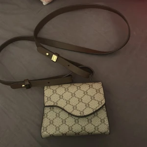 Mönstrad axelväska med brunt läderband - Snygg axelväska med ett klassiskt mönster i beige och brunt. Väskan har en justerbar axelrem i brunt läder och en stilren design. Perfekt för både vardag och festliga tillfällen!