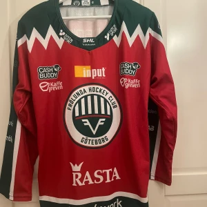 Hockeytröja från Frölunda HC - Säljer en officiell Frölunda HC hockeytröja för säsongen 23/24. Tröjan är röd med gröna detaljer och har flera sponsormärken. Inte ens provad, så helt ny utan prislapp. Perfekt för att heja på laget i stil! 🏒