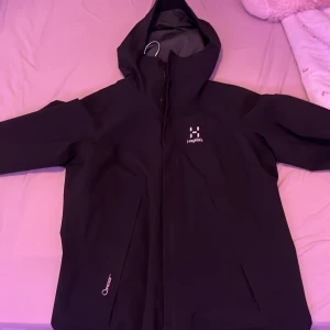 Svart regnjacka från Haglöfs - Säljer en svart regnjacka från Haglöfs i storlek S. Jackan har en praktisk huva och dragkedja framtill. Tillverkad i vattentätt material med Q Proof-teknologi för extra skydd mot regn. Perfekt för blöta dagar!