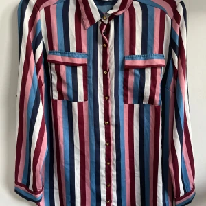 Randig skjorta från Sincerely Loved - Snygg randig skjorta i blått, vinrött, rosa och vitt från Sincerely Loved. Skjortan har långa ärmar och knappar framtill. Perfekt för både vardag och festliga tillfällen. Den är i nytt skick och redo att bäras med stil!