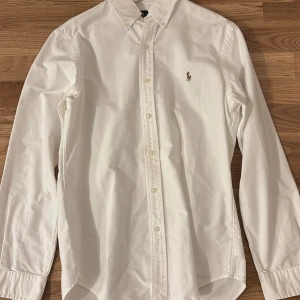 Vit skjorta från Ralph Lauren - Skick 9/10 knappt använd. Slim fit vit skjorta från Ralph Lauren med broderad logotyp på bröstet. Skjortan har långa ärmar och knappar framtill. Perfekt för en stilren look.