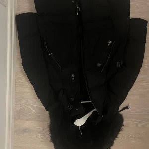 Svart dunjacka från Cedrico - Snygg svart dunjacka från Cedrico med pälskantad huva och dragkedja. Perfekt för kalla dagar med sin varma och skyddande design. Jackan har flera praktiska fickor med dragkedjor.