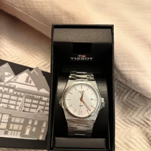 Super snygg herr klocka från Tissot - Elegant silverfärgad klocka från Tissot med vit urtavla och datumvisning. Klockan har ett stilrent armband i rostfritt stål och tydliga markeringar. Perfekt för den som söker en klassisk och tidlös design. Aldrig använd med plasten kvar, alla tillbehör med kommer. 