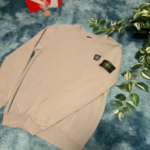 Beige sweatshirt från Stone Island - Säljer en stilren beige sweatshirt från Stone Island med deras ikoniska märke på ärmen. Tröjan har en rund halsringning och ribbade muddar vid ärmslut och nederkant. Perfekt för en avslappnad look.
