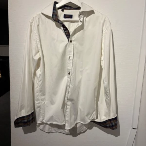 Vit skjorta med rutiga detaljer - Snygg vit skjorta med unika rutiga detaljer på insidan av kragen och ärmsluten. Perfekt för en stilren look med en twist. Skjortan har knappar framtill och långa ärmar.