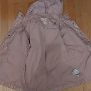 Rosa regnjacka från Zara  - Säljer en snygg rosa regnjacka från Zara med luva och knappar. Perfekt för regniga dagar och håller dig torr med stil. Jackan har en glansig finish och är designad för att vara både praktisk och trendig.