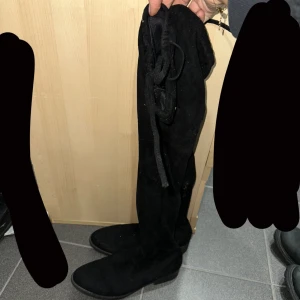 Svarta overknee stövlar - Snygga svarta overknee stövlar i mocka med snörning baktill. Perfekta för höst och vinter, dessa stövlar ger en stilren look och håller dig varm. De har en rund tå och en låg klack för bekväm användning hela dagen.