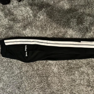 Palm Angels byxor - Säljer ett par svarta byxor från Palm Angels med vita ränder längs sidorna. De har en elastisk midja och en avslappnad passform. Perfekta för en casual look eller träning. Byxorna är i mycket bra skick och redo för nya äventyr!