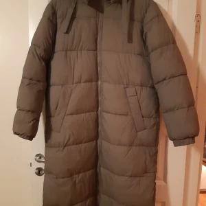 Lâng dunjacka - Säljer en lång dunjacka i mycket bra skick. Den är perfekt för kalla vinterdagar med sin quiltade design och långa ärmar. Jackan har en huva och stängs med dragkedja. Supermysig och varm!