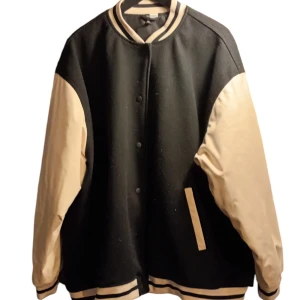 Svart och beige bomberjacka - Snygg oversize svart och beige bomber/baseballjacka med kontrasterande ärmar och ribbade muddar. Jackan har knappar framtill och är tillverkad i 100% polyester. Perfekt för en cool och avslappnad stil.