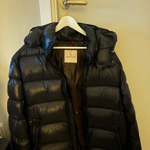 Svart dunjacka från Moncler - Snygg svart dunjacka från Moncler med glansig finish och huva. Jackan har en dragkedja framtill och är perfekt för kyligare väder. Den är långärmad och har en klassisk pufferdesign.