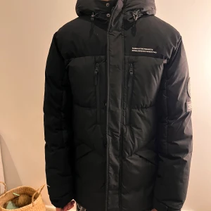 Svart dunjacka från Black Squad - Säljer en svart dunjacka från Black Squad med huva och flera praktiska fickor. Jackan har en stilren design med broderade detaljer på ärmen och bröstet. Perfekt för kalla dagar.