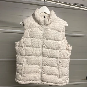 Vit quiltad dunväst - Säljer en snygg vit quiltad dunväst med diskret mönster. Västen har en dragkedja framtill och är perfekt för kyliga dagar. Den är både stilren och praktisk, idealisk för lager-på-lager under höst och vår. Säljes i befintligt skick. Storlek L