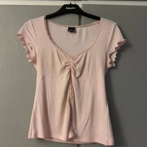 Ljusrosa topp från Gina Tricot - Söt ljusrosa topp från Gina Tricot i storlek S. Den har korta ärmar med volangdetaljer och en v-ringad design med spetskant. Toppen har en snygg rynkning framtill som ger en fin passform. Perfekt för vår och sommar!
