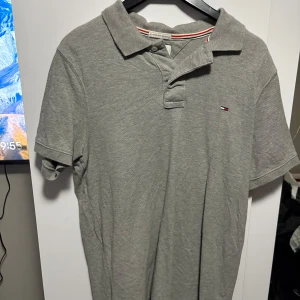 Grå pikétröja från Tommy Hilfiger - Snygg grå pikétröja från Tommy Hilfiger med klassisk krage och knappslå. Den har en liten broderad logga på bröstet och ribbade ärmslut. Perfekt för en avslappnad stil.