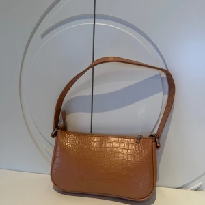 Orange/brun axelväska med krokodilmönster - Säljer en snygg orange/brun axelväska i syntetmaterial med krokodilmönster. Väskan har en dragkedja upptill, ett fack med dragkedja inne i väskan och två mindre fack. Perfekt för både vardag och fest! 👜