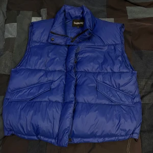 Blå dunväst från b.young - Säljer en snygg blå dunväst från b.young. Västen har en hög krage och stängs med dragkedja och knappar. Perfekt för kyliga dagar och ger en stilren look. Passar bra till lager-på-lager-stil.