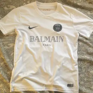 Säljer en snygg vit fotbollströja i samarbete mellan Nike och Balmain. Tröjan har korta ärmar och är i Dri-FIT-material för bästa komfort. Perfekt för både träning och vardag. Den har en stilren design med Balmain Paris-logga på bröstet. Passa på att köpa denna unika tröja!   Den är i super bra skick!!