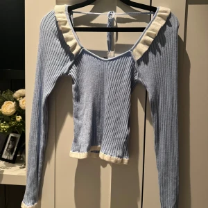 Ljusblå ribbad topp med volangdetaljer - Superfin ljusblå ribbad topp med vita volangdetaljer runt halsen. Den har långa ärmar och en söt knytning i ryggen. Perfekt för vår och sommar! 🌸