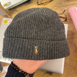 Grå ribbad mössa från Polo Ralph Lauren - Snygg grå ribbad mössa från Polo Ralph Lauren i 100% merinoull. Den har en broderad logga framtill och är perfekt för kyliga dagar. Mössan är i mycket bra skick och passar till både vardag och lite finare tillfällen. Ny skick 