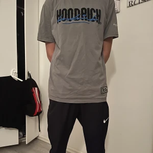 Grå t-shirt från Hoodrich - Säljer en grå t-shirt från Hoodrich med svart och blå logga på framsidan. T-shirten har en avslappnad passform och korta ärmar. Perfekt för en casual look.
