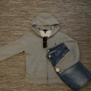 Grå hoodie från Ralph Lauren - Säljer en snygg och bekväm grå hoodie från Polo Ralph Lauren. Den har en dragkedja framtill och en klassisk huva. Perfekt för en avslappnad stil och passar bra till jeans. Den är i mycket bra skick och redo för nya äventyr! Sällan använd då jag inte tyckte den passade min stil. Storleken är från 18-20 vilket är som S.