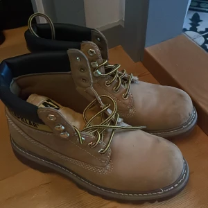 Beige boots från Caterpillar - Säljer ett par robusta och snygga beige boots från Caterpillar. De har en klassisk design med snörning och en slitstark sula. Perfekta för höst och vinter, och de är i bra skick. Skorna har en bekväm passform och är gjorda i skinn med en vadderad krage för extra komfort.