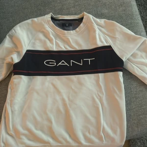 Vit tröja från GANT - Säljer en snygg vit tröja från GANT med ett marin blått band över bröstet där märket står i vitt. Tröjan har långa ärmar och en rund halsringning. Perfekt för en casual look eller en avslappnad dag ute. 🖤