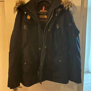 Svart parkajacka med pälsdetaljer - Säljer en svart parkajacka med pälsdetaljer på huvan. Jackan har dragkedja och knappar framtill, perfekt för kalla vinterdagar. Den är långärmad och har en klassisk stil som passar både till vardags och fest. Huvan är avtagbar och jackan har flera praktiska fickor.