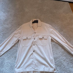 Beige långärmad skjorta med knappar - Säljer en stilren beige skjorta med långa ärmar och knappar framtill. Skjortan har två bröstfickor med knappdetaljer. Perfekt för både vardag och festliga tillfällen. Passar bra till höst och vår.