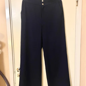 Byxor från Holly & White - Snygga mörkblåa byxor från Holly & White i storlek 38. De har en hög midja och vida ben, vilket ger en bekväm och stilren look. Byxorna har dekorativa knappar framtill.