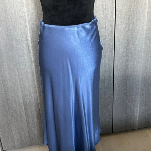 Blå långkjol i satin - Säljer en elegant blå midikjol i satin. Kjolen har en slät och glansig yta som ger en lyxig känsla. Perfekt för festliga tillfällen eller en kväll ute. Den har en enkel och stilren design som gör den lätt att matcha med olika toppar.