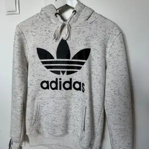 Säljer en snygg gråmelerad hoodie från Adidas med det klassiska logotyptrycket i svart på framsidan. Den har en stor ficka fram och justerbar huva med snören. Perfekt för en avslappnad stil eller träning. Superbekväm och i bra skick!