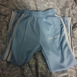 Ljusblå shorts från Palm Angels - Säljer ett par ljusblå shorts från Palm Angels. De har en elastisk midja med snörning och vita ränder längs sidorna. Perfekta för en avslappnad stil eller träning. Märkesloggan syns tydligt på sidan.