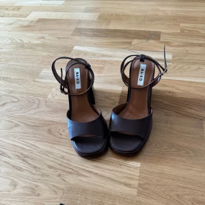 Bruna klackskor från A.K.D - Säljer ett par snygga bruna klackskor från na-kd De har en bred rem över tårna och en justerbar rem runt ankeln. Klackarna är höga och stabila, vilket gör dem perfekta för både fest och vardag. Skorna är i skinn och har en elegant design.