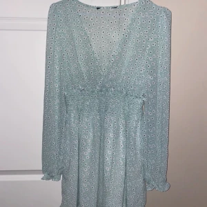 Ljusblå blommig klänning - Säljer en superfin ljusblå klänning med blommigt mönster. Den har en v-ringad design och långa ärmar